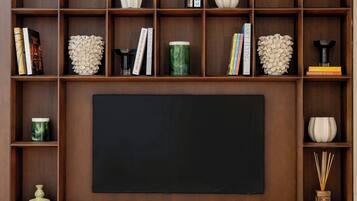 A living room with a large flat-screen TV, wooden shelving with books and decorative items, a white armchair, and a small round table.