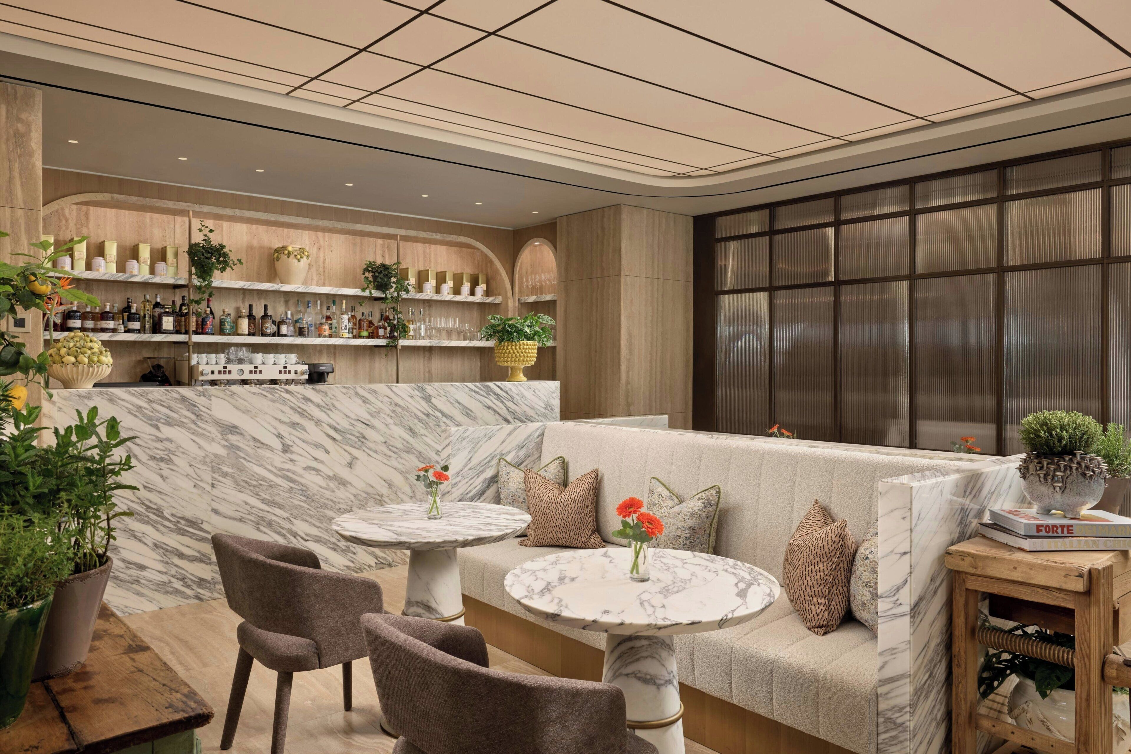 A bar area with shelves of bottles and glasses, a marble wall, a seating area with a sofa and round tables, and a large window with a view.