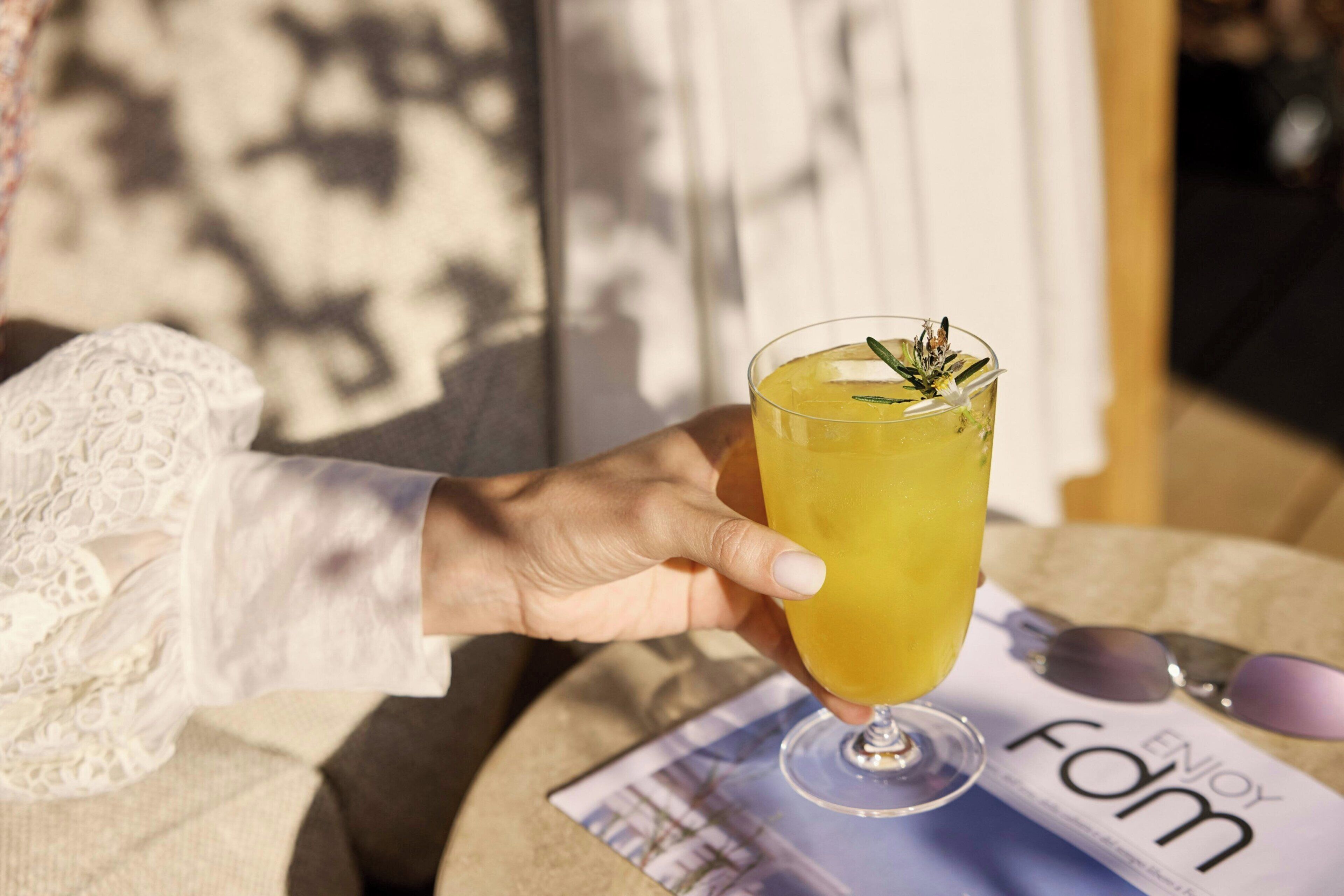Eine Hand hält ein Glas mit einem gelben Getränk, ein Magazin mit der Aufschrift 'ENJOY FOM' und eine Sonnenbrille liegen auf einem Tisch.