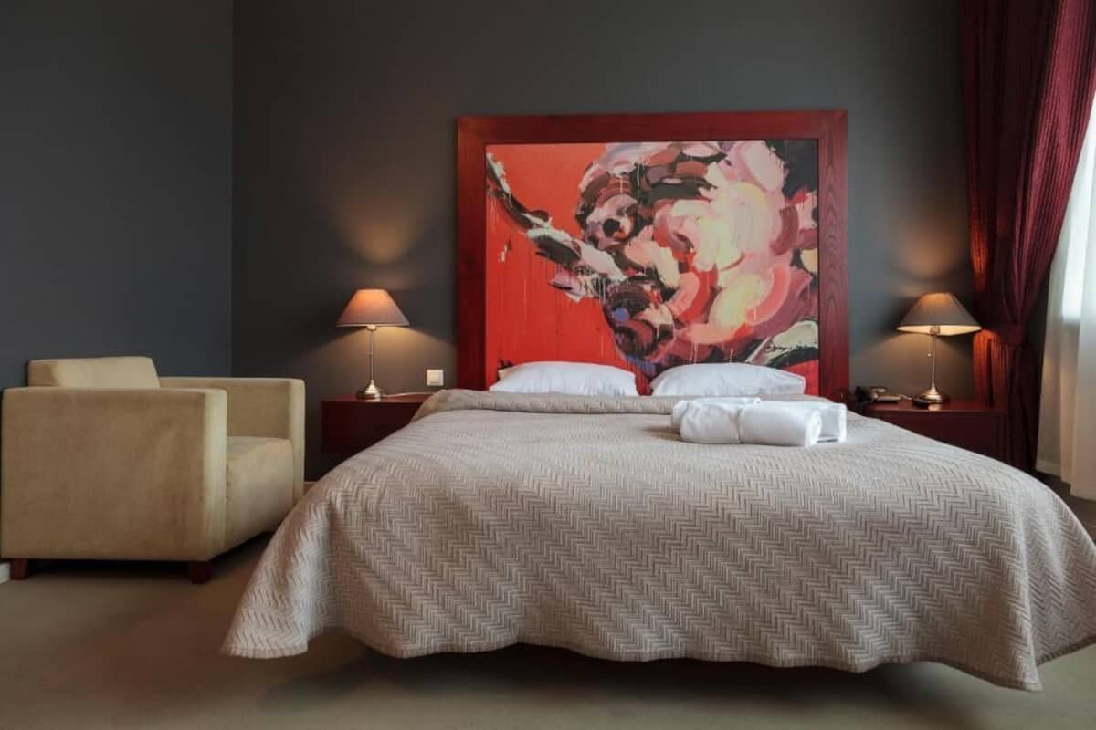A hotel room with a large bed, two bedside tables with lamps, a chair, and a large abstract painting on the wall.
