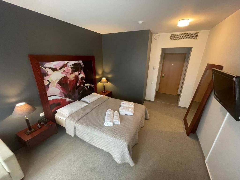 Habitación de hotel con una cama, dos mesitas de noche, un cuadro abstracto grande, una puerta y un televisor.