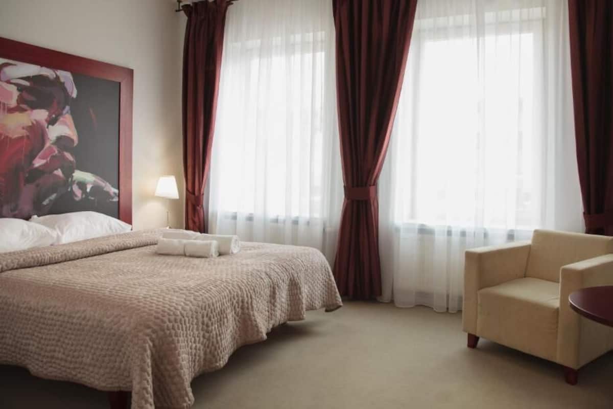 A hotel room with a large bed, a chair, a small table, and a painting on the wall.