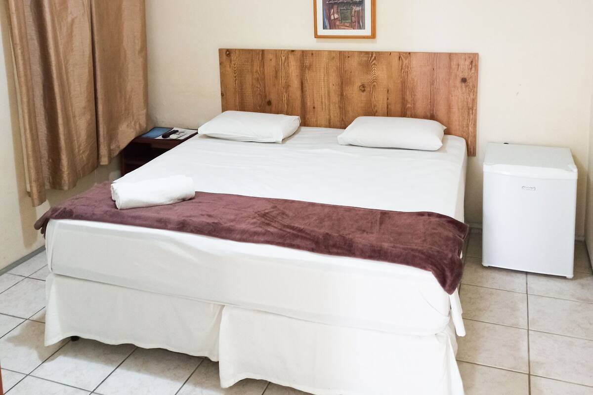 A hotel room with a wooden headboard, a bed with white linens, a small bedside table, a white refrigerator, and a window with curtains.