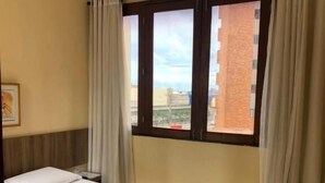 Habitación de hotel con cama, mesita de noche, ventana con cortinas y vista a edificios.
