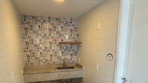 Una pequeña cocina con salpicadero de azulejos, una encimera, un lavamanos, un cubo de basura negro y una puerta.