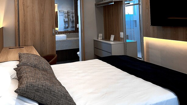 Habitación de hotel moderna con una cama grande, un armario de madera, televisión empotrada y un baño visible a través de una puerta abierta.