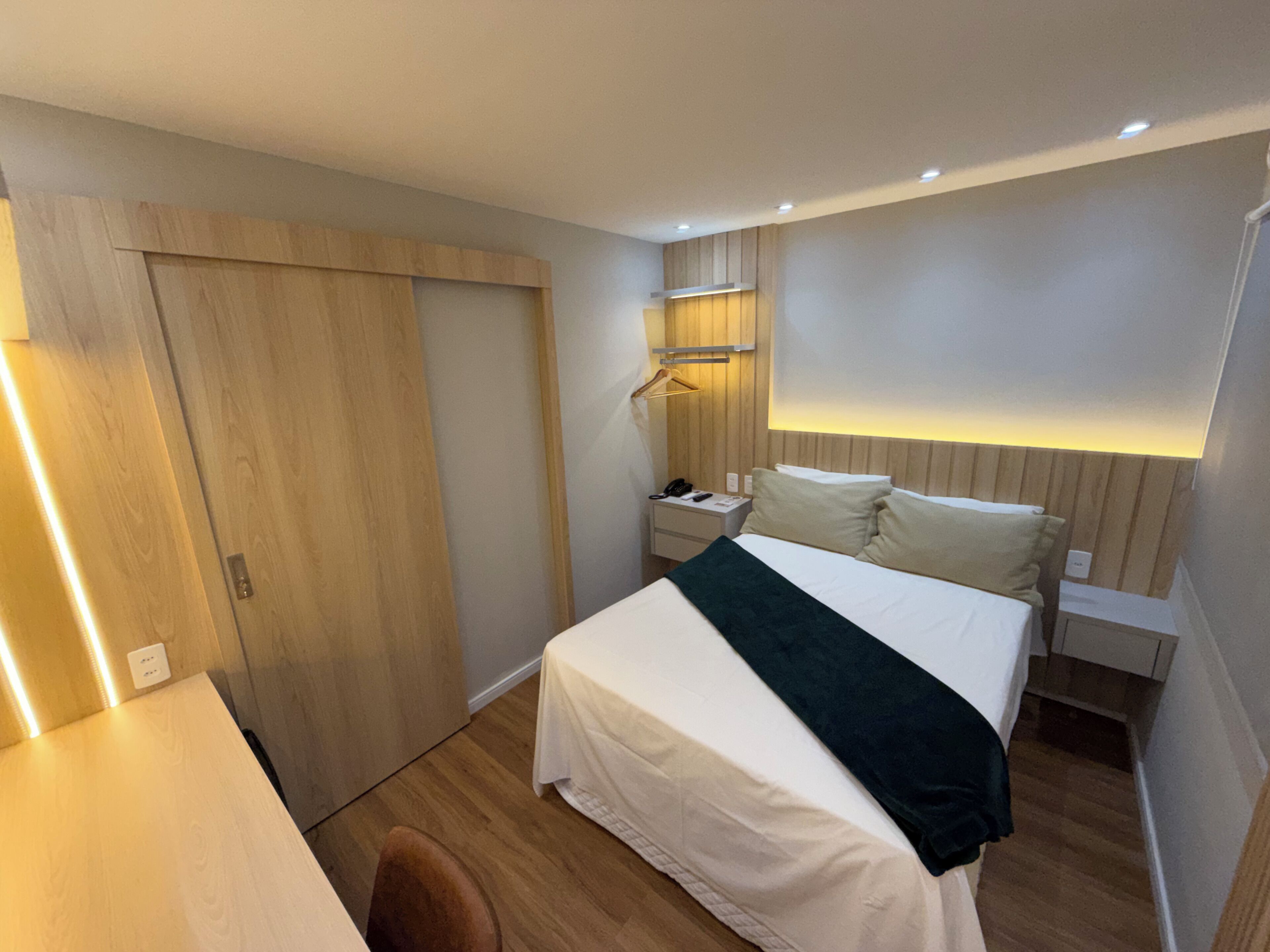 Una habitación de hotel con un armario de madera, una mesita de noche con lámpara, una cama con ropa blanca y un aplique de pared.