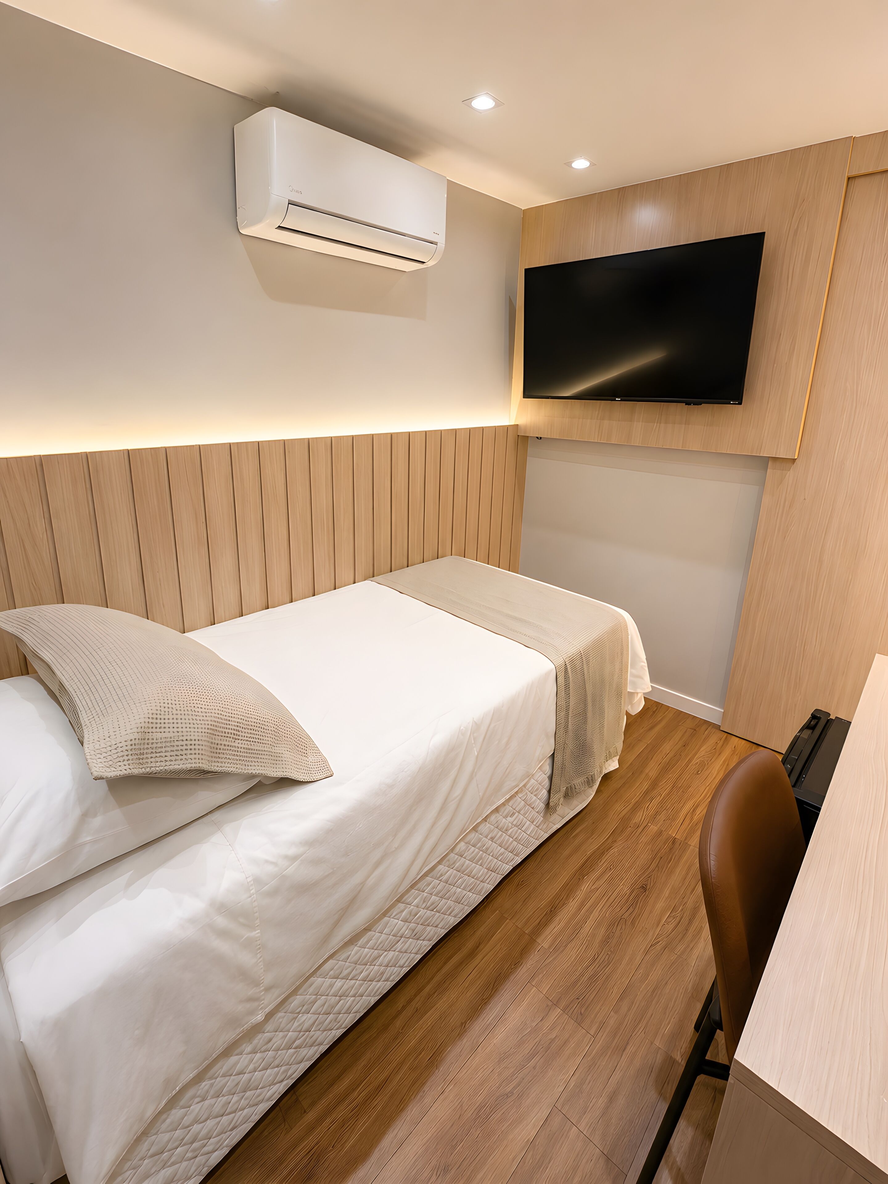 Une chambre d’hôtel moderne équipée d’un lit, d’un bureau, d’une chaise, d’un climatiseur et d’un téléviseur fixé au mur.