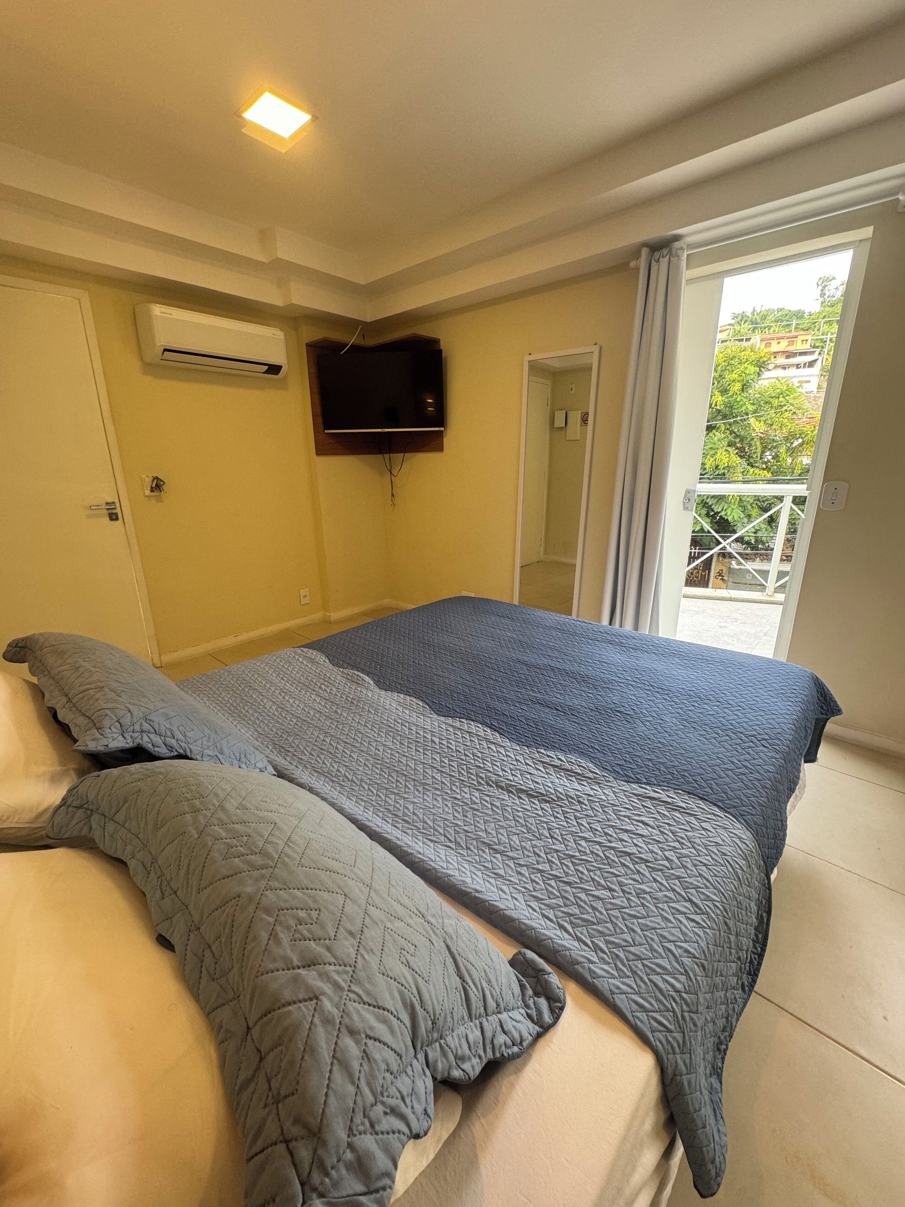 A bedroom with a bed, a flat-screen TV mounted on the wall, and a window with a view of greenery outside.