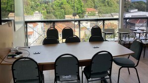 Un salón de conferencias con una gran mesa y sillas, ventanas amplias que dan a vistas de edificios y árboles, y un televisor montado en la pared.