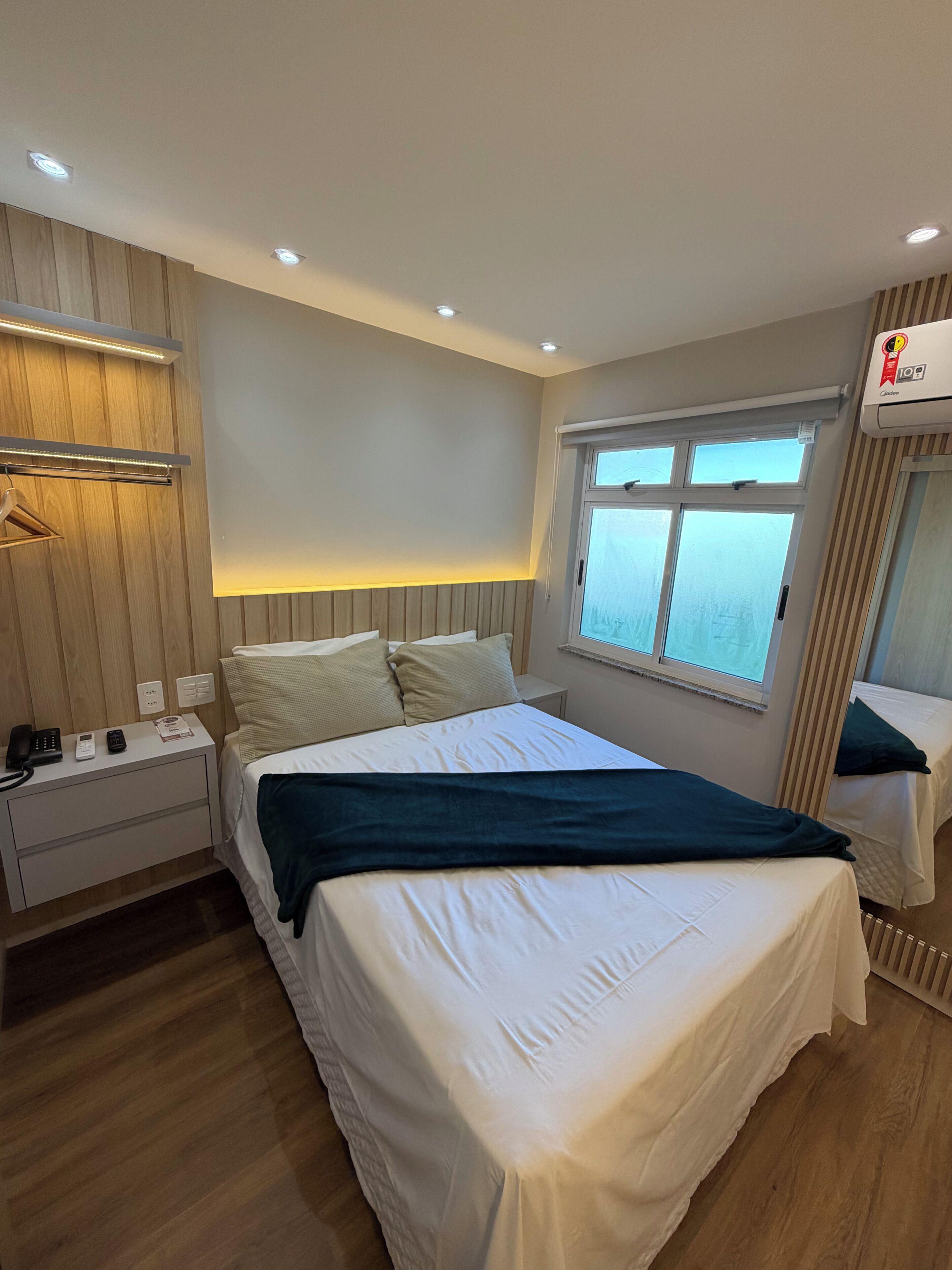 Habitación de hotel con cama, mesitas de noche, perchero, ventana con vistas y aire acondicionado.