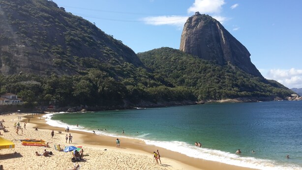 Uma praia com águas turquesa cristalinas, uma grande formação rochosa e uma orla de areia com pessoas e guarda-sóis.