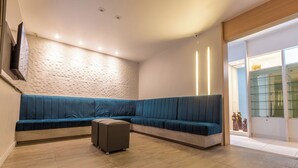 A modern living room with a blue sectional sofa, a small ottoman, and a flat-screen TV mounted on the wall. - Rede Andrade Navegantes (Recife)