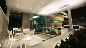 A modern outdoor dining area with white chairs and a glass table, overlooking a cityscape at night. - Rede Andrade Navegantes (Recife)