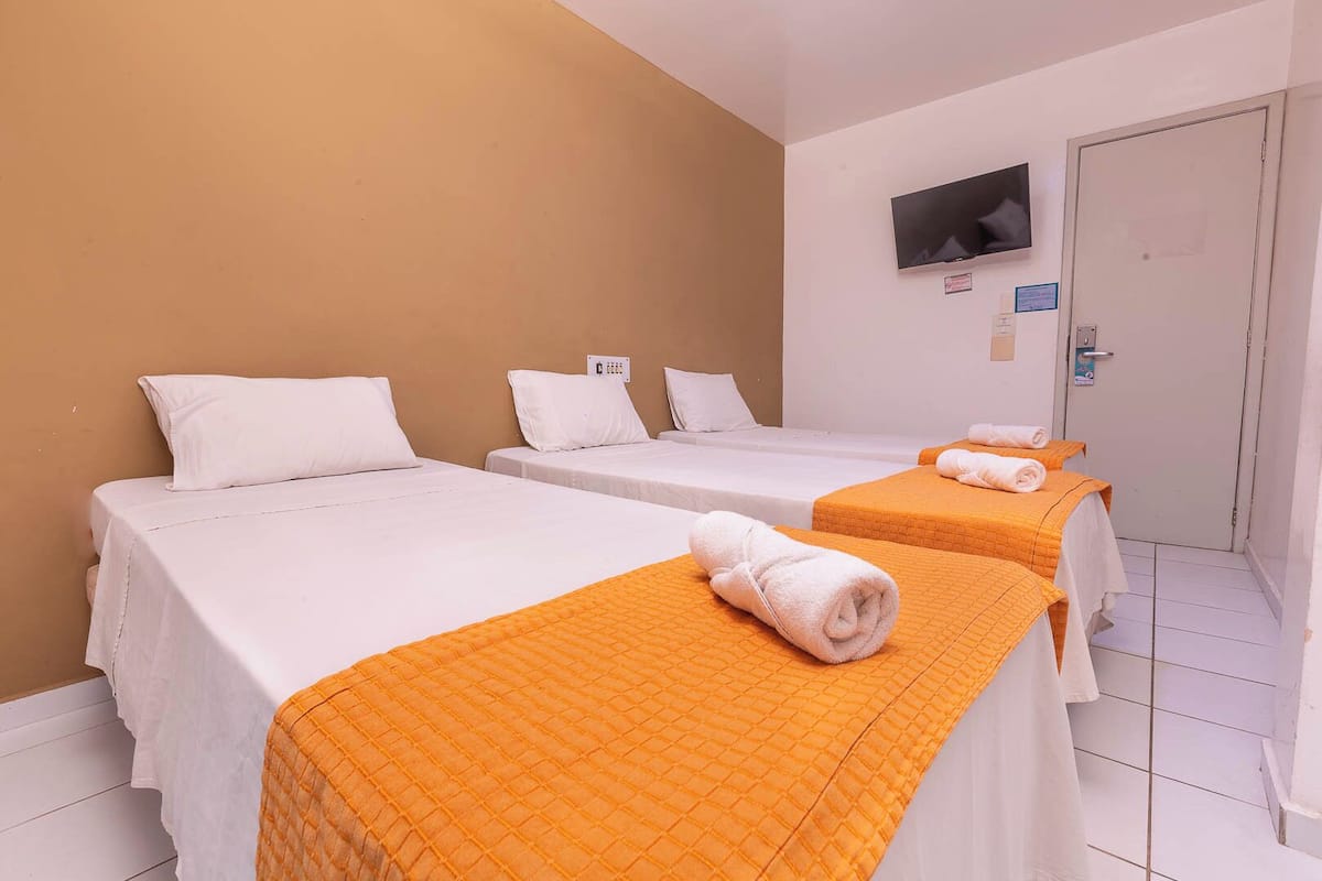 A hotel room with three single beds, each with white bedding and an orange quilted cover. There is a flat-screen TV mounted on the wall, and a door with a sign on it.