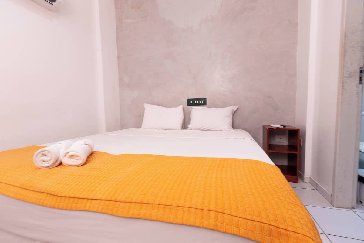 A single bed with a white mattress and two pillows, a yellow quilt, and rolled towels on an orange bedspread.