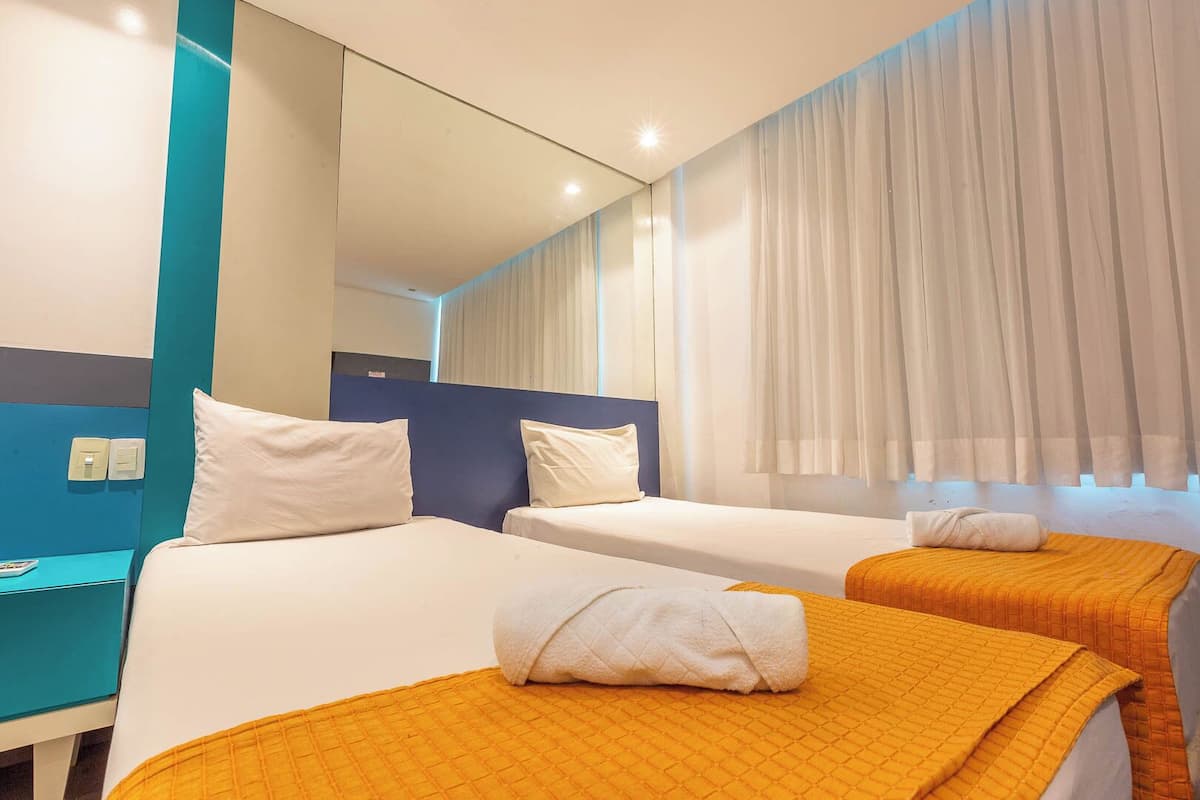 A hotel room with two beds, each with a white pillow and an orange blanket. There is a mirror on the wall and a window with white curtains.