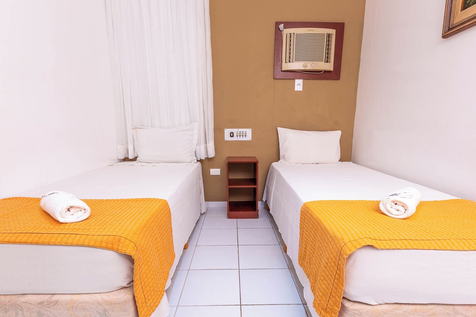 A hotel room with two single beds, each with a yellow blanket and a rolled towel. There is a small wooden shelf between the beds, and a wall-mounted heater above the shelf.