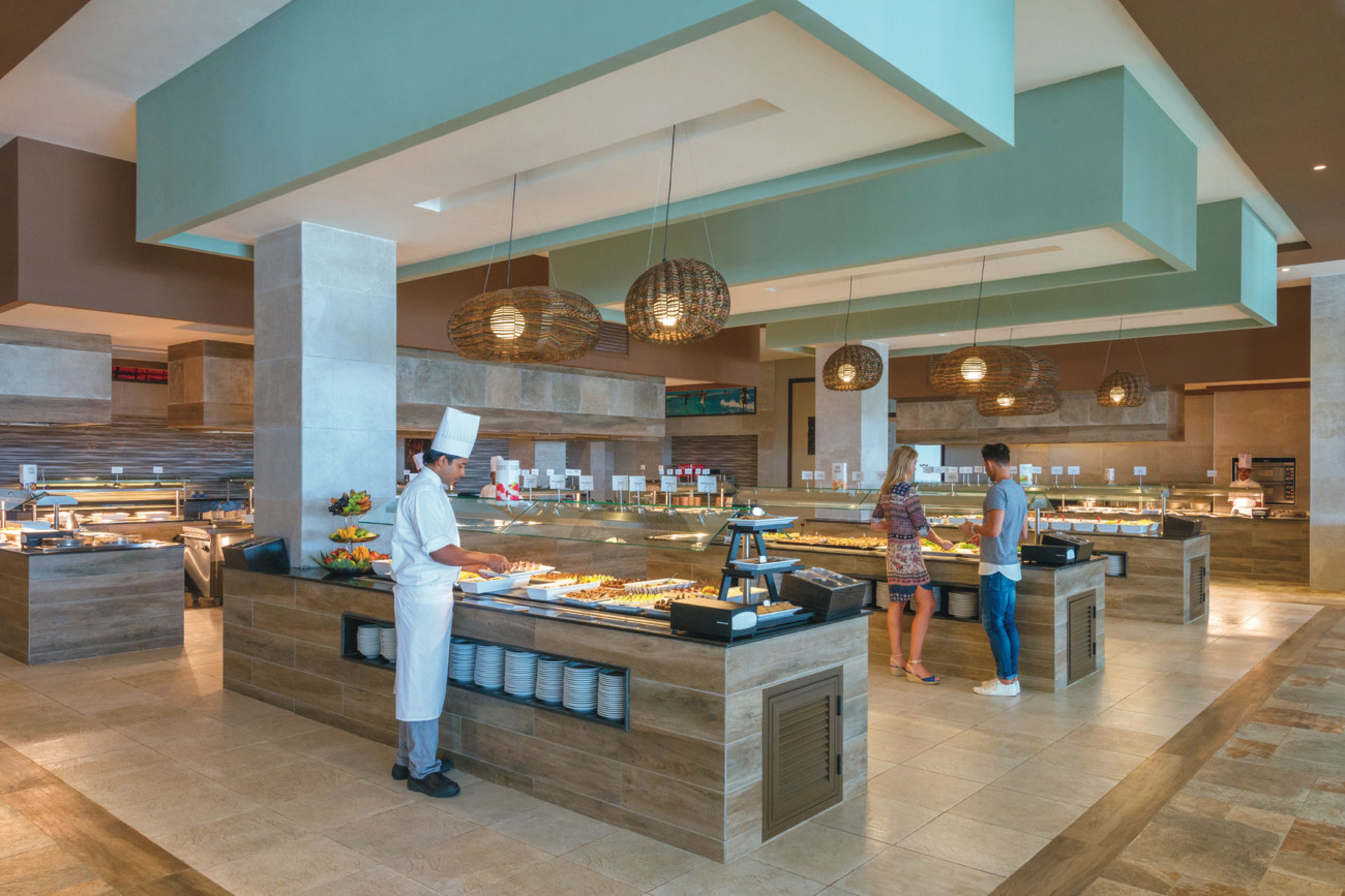 Zona de bufé con diversas opciones de comida, un miembro del personal atendiendo a los huéspedes y un diseño interior moderno con luces colgantes.