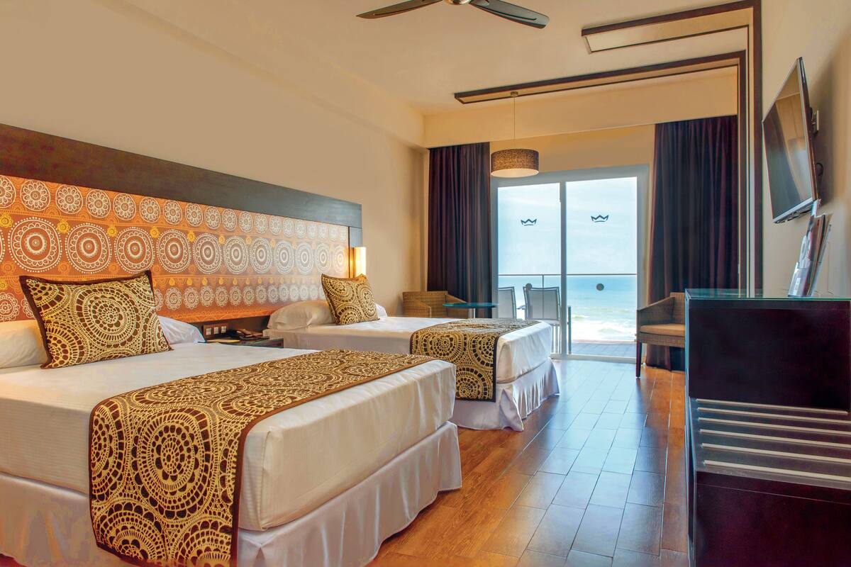 A hotel room with two beds, a desk, a TV, and a view of the ocean.