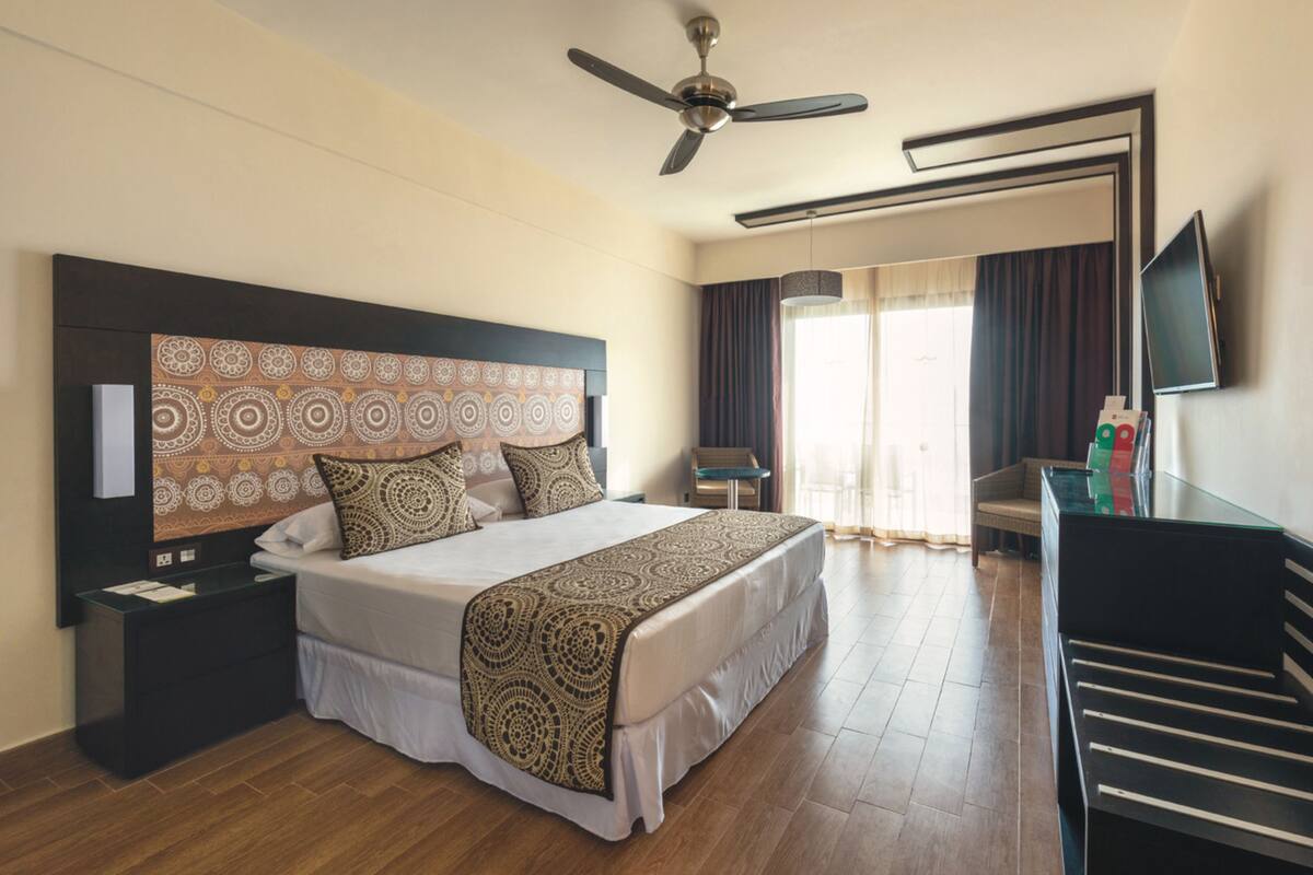 A hotel room with a large bed, a ceiling fan, a TV, and a piano.