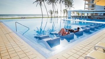 Une piscine avec des marches permettant d’y accéder, entourée de palmiers et offrant une vue sur l’océan.