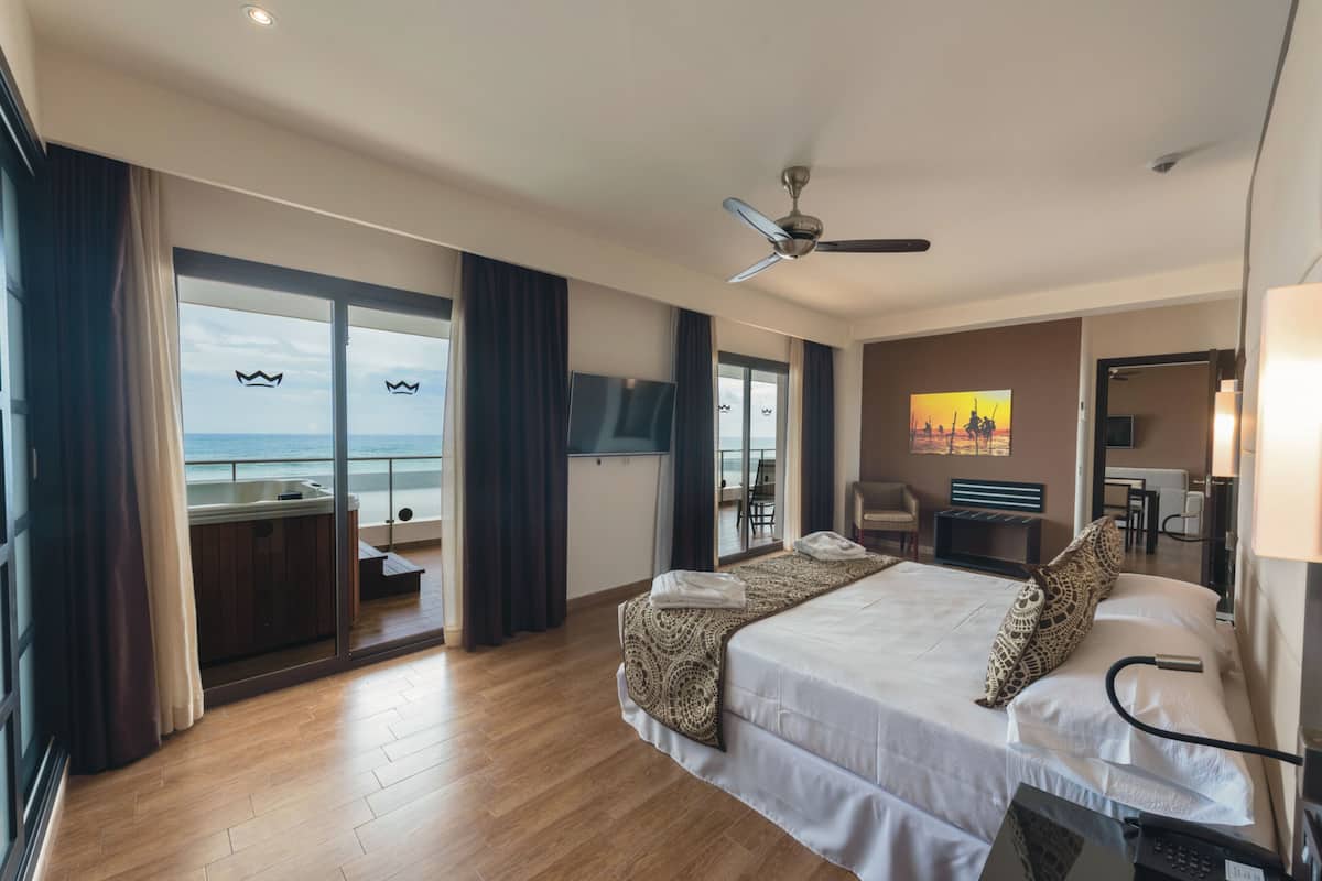 A hotel room with a large bed, a ceiling fan, a TV, a desk, and a view of the ocean through a sliding door.