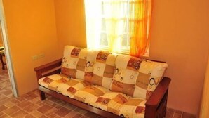 A cozy room with a wooden sofa against a wall, a window with curtains, and a tiled floor. - Angel Fish Cove (Buccoo)