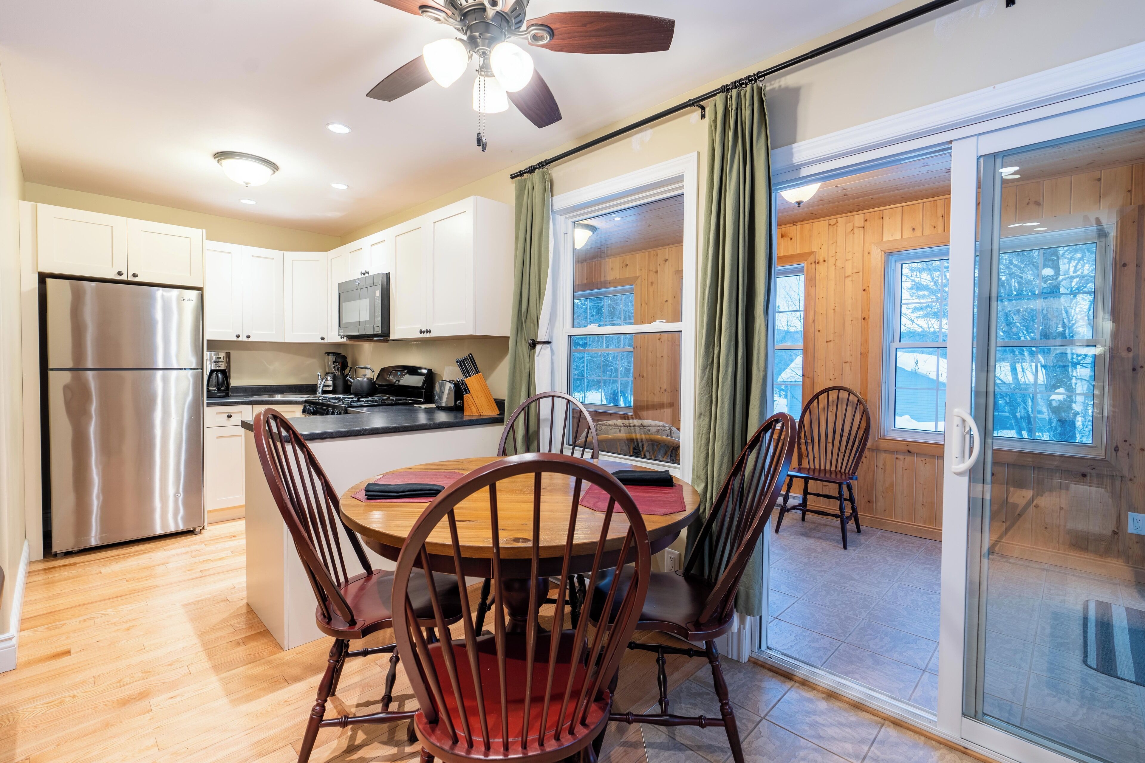 A modern kitchen with a stainless steel refrigerator, wooden cabinets, a dining table with chairs, and a large window with curtains.