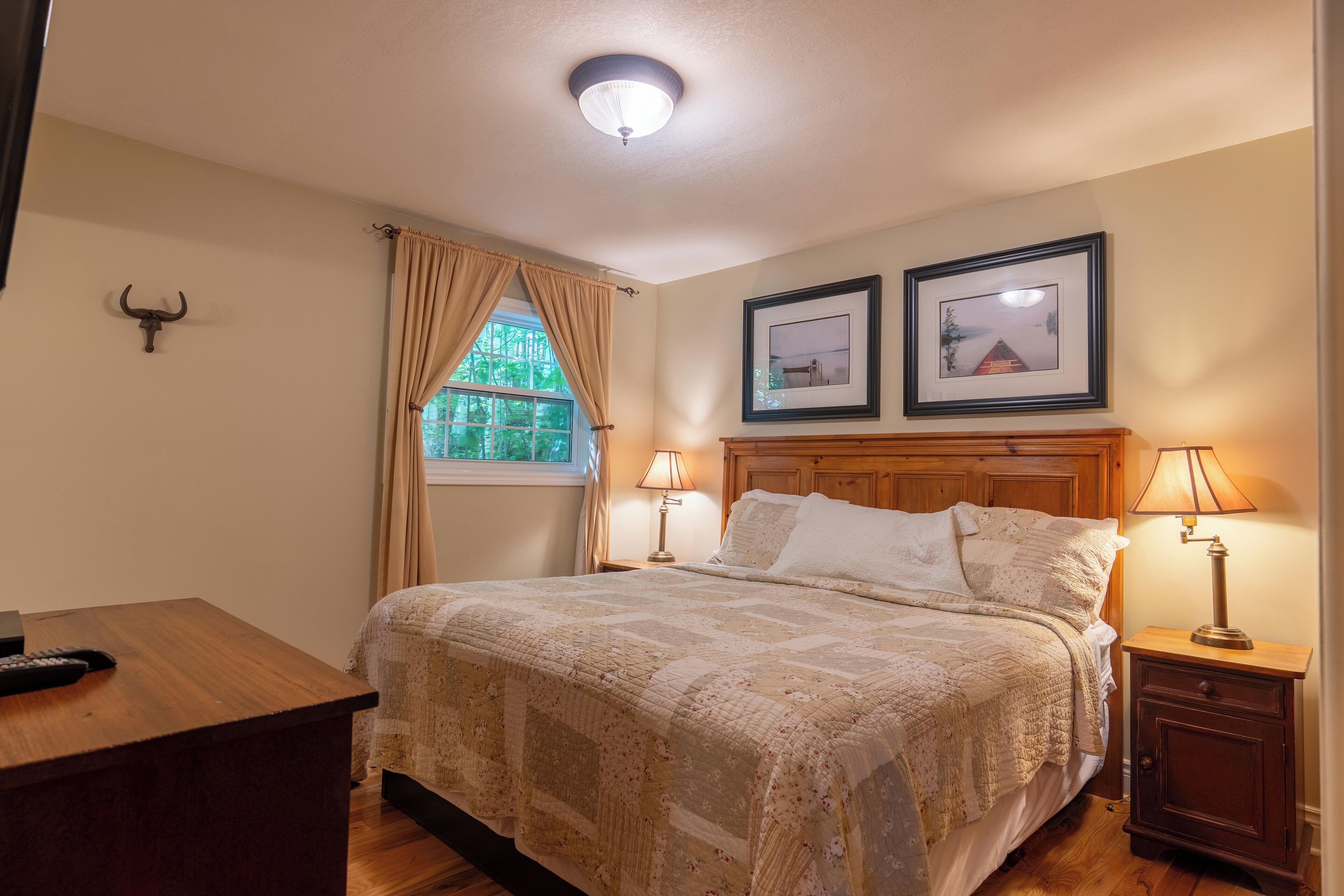 Une chambre à coucher avec un grand lit, deux lampes de chevet, une table de nuit en bois, un bureau, une télécommande, une décoration représentant une tête de taureau et deux tableaux encadrés accrochés au mur.