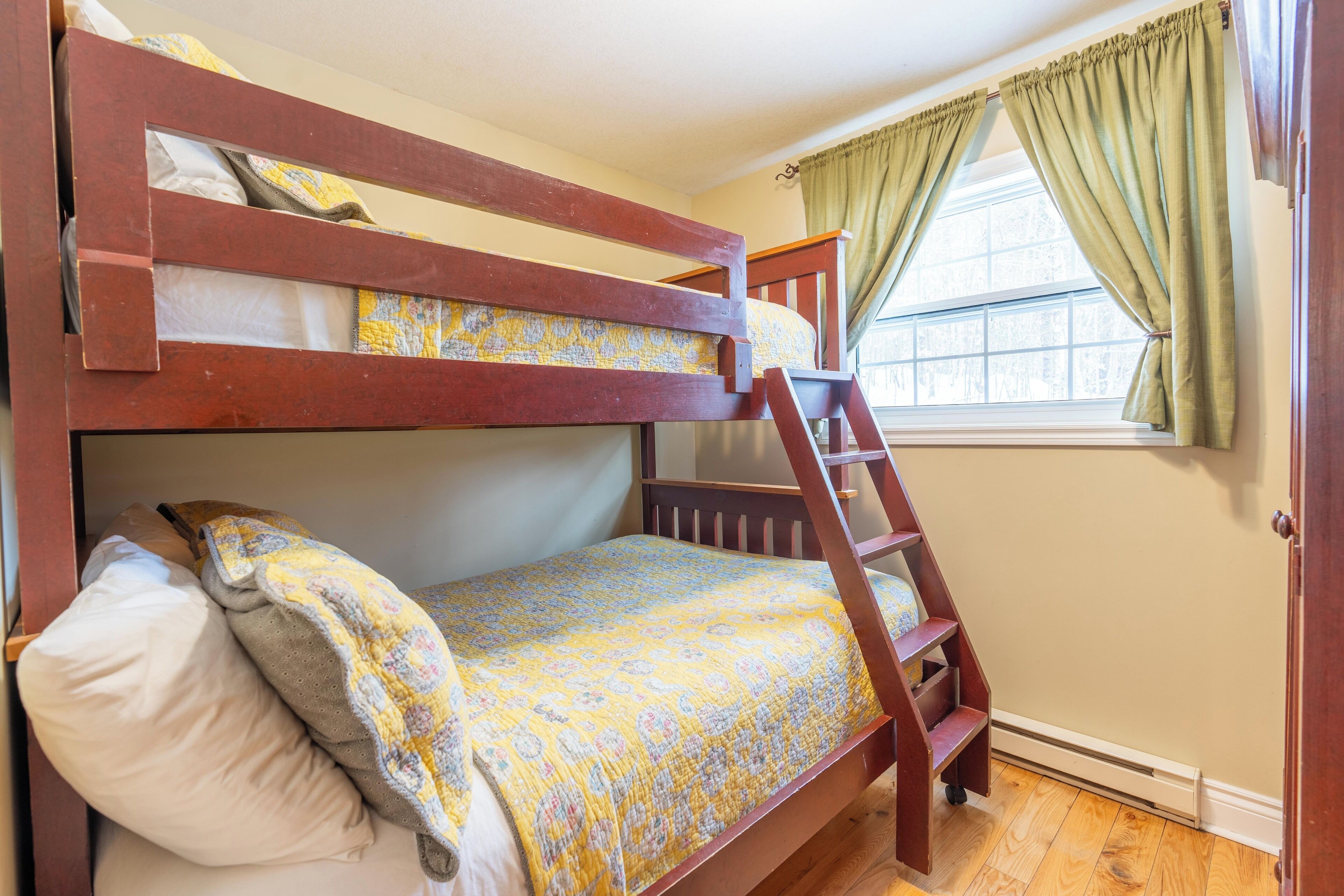 A bunk bed with a ladder, a window with curtains, and a wooden floor.