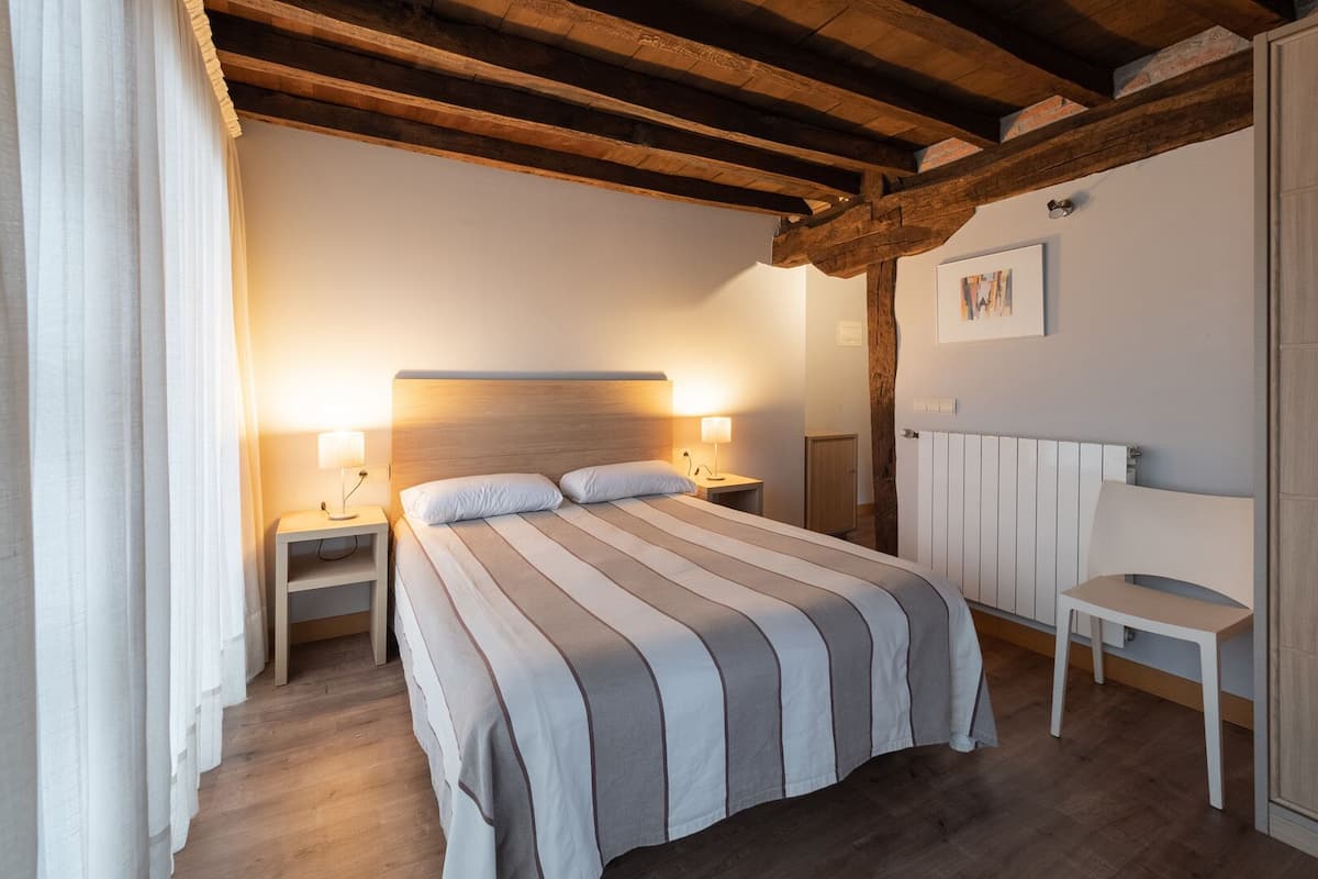 Un dormitorio con techo de madera, una cama con ropa de cama a rayas, lámparas de noche, una silla y un radiador.