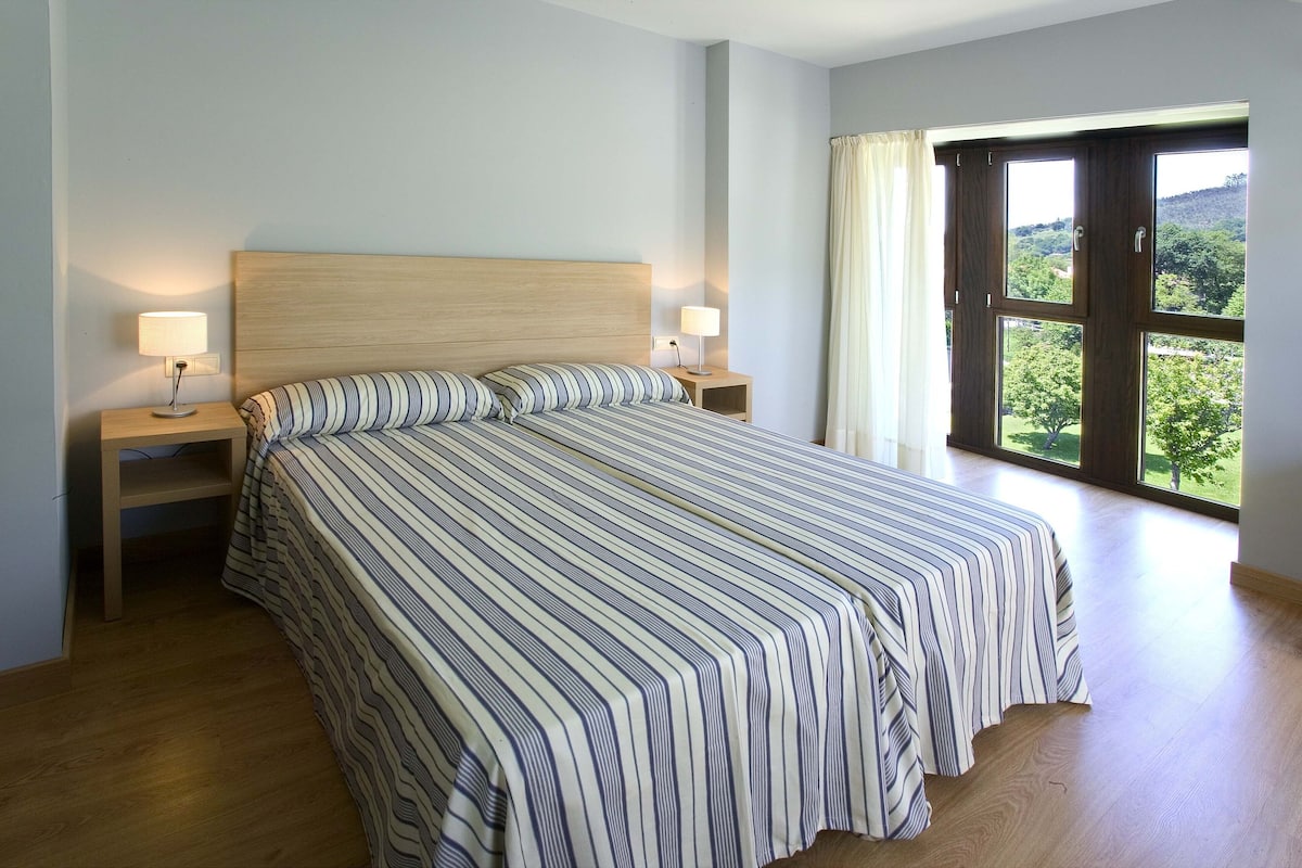 Un dormitorio con una cama grande, dos mesitas de noche con lámparas, un cabecero de madera y un ventanal con vistas a la vegetación.