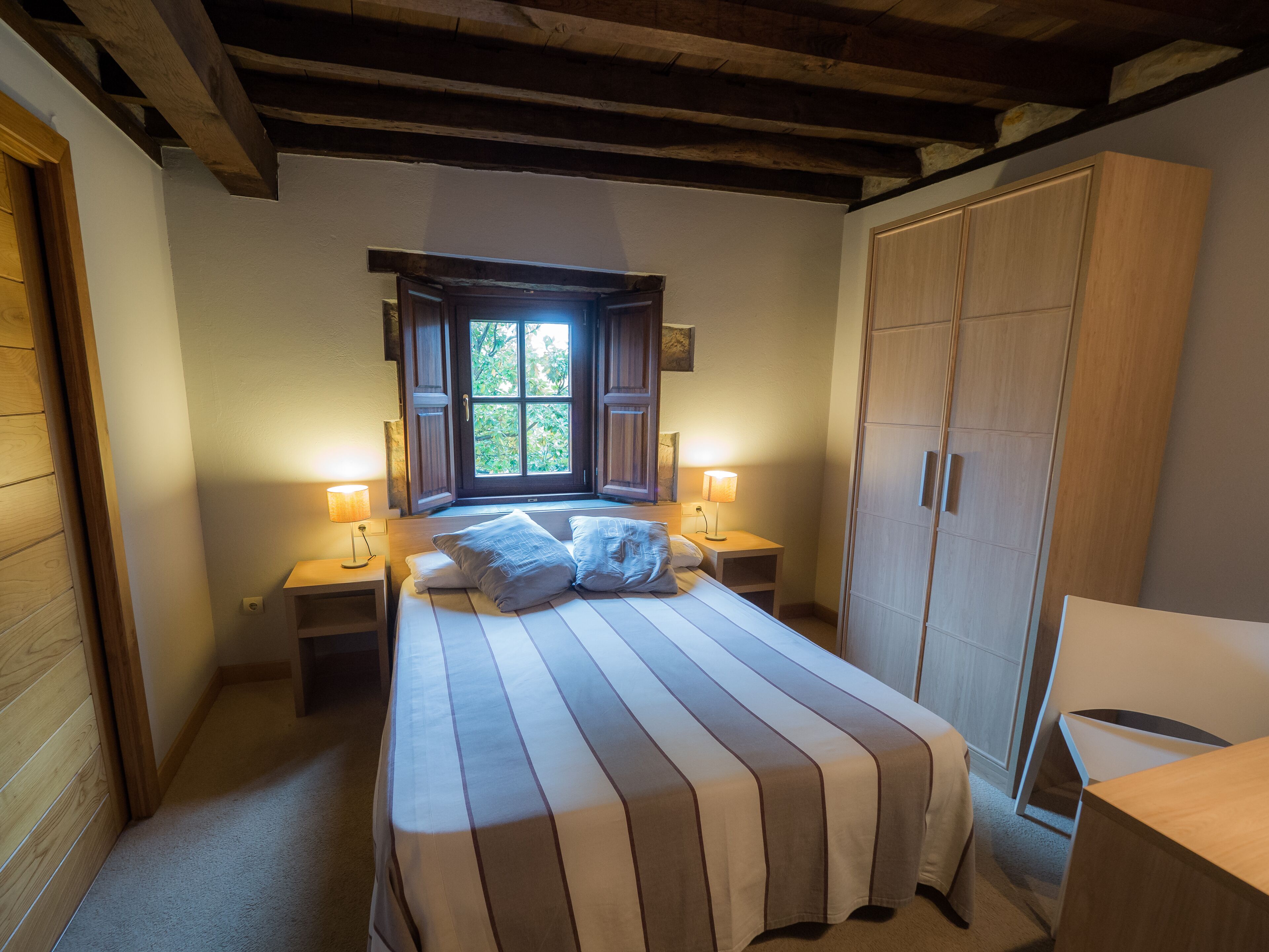 A bedroom with a large bed, two bedside tables with lamps, a window with a view of greenery, and a wardrobe.