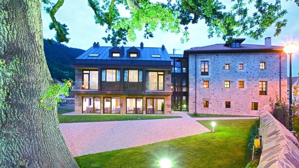 A modern building with a glass facade and a stone exterior, featuring a courtyard with a lawn and a large tree. - Apartamentos Posada La Robla (Castaneda)