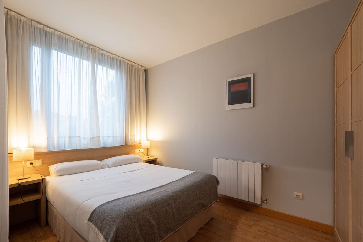 Habitación de hotel con cama, lámparas de noche, radiador y ventana con cortinas traslúcidas.