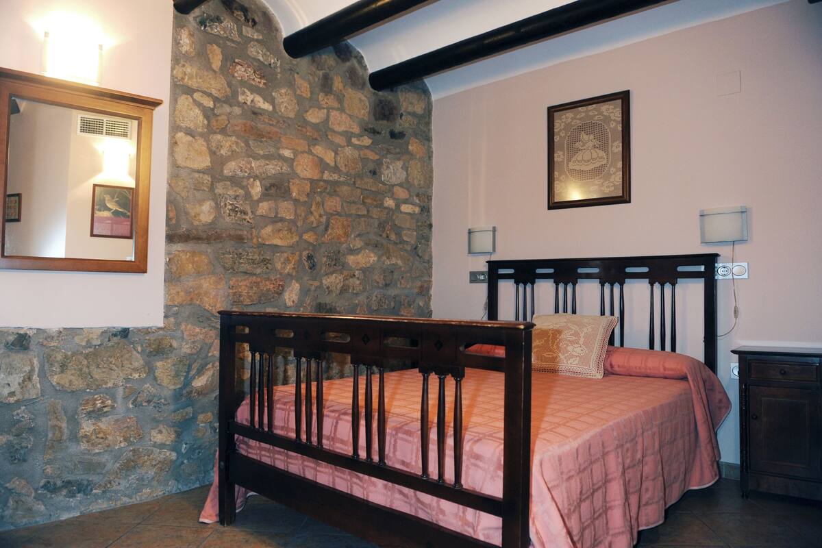 A bedroom with a stone wall, a wooden bed with a pink bedspread, a mirror, and two wall-mounted lamps.