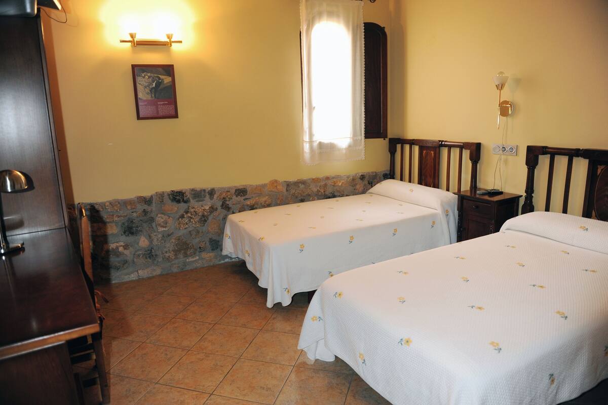 A room with two beds, a wooden cabinet, a stone wall, and a window with a curtain.