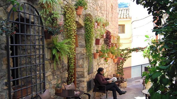 Un hombre leyendo un periódico en un área de estar al aire libre con paredes de piedra, plantas en macetas y una puerta de metal.
