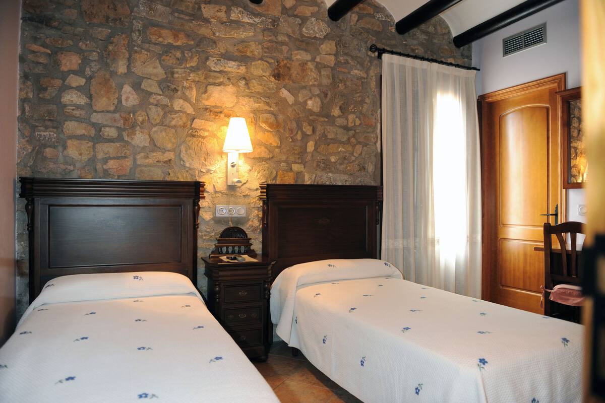 A room with two beds, a stone wall, a wooden door, and a nightstand with a lamp.