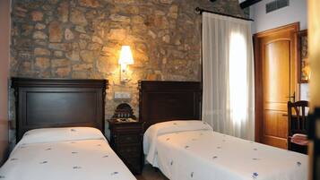 A room with two beds, a stone wall, a wooden door, and a nightstand with a lamp.