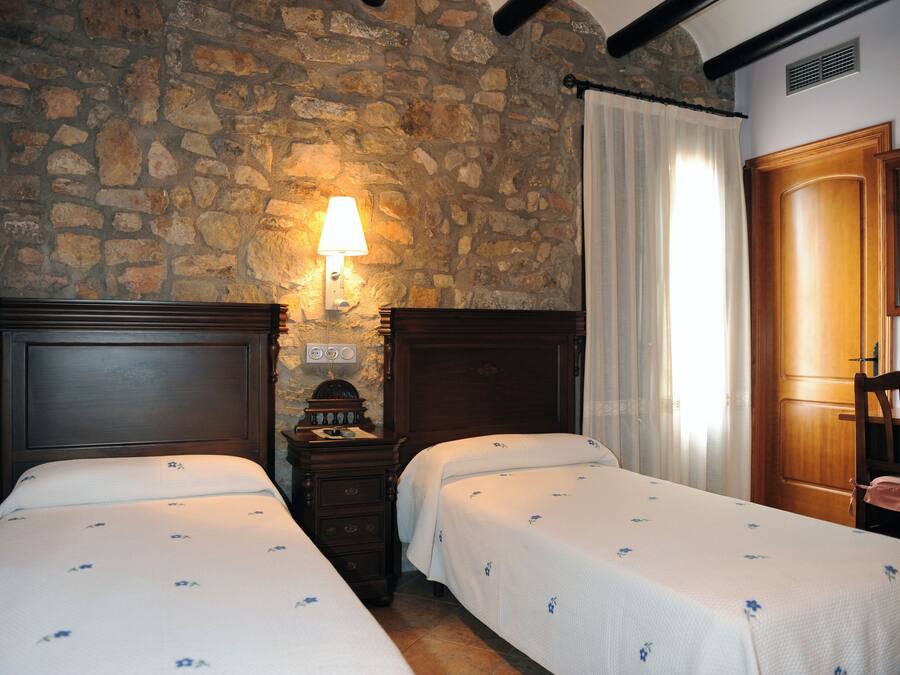 A room with two beds, a stone wall, a wooden door, and a nightstand with a lamp.