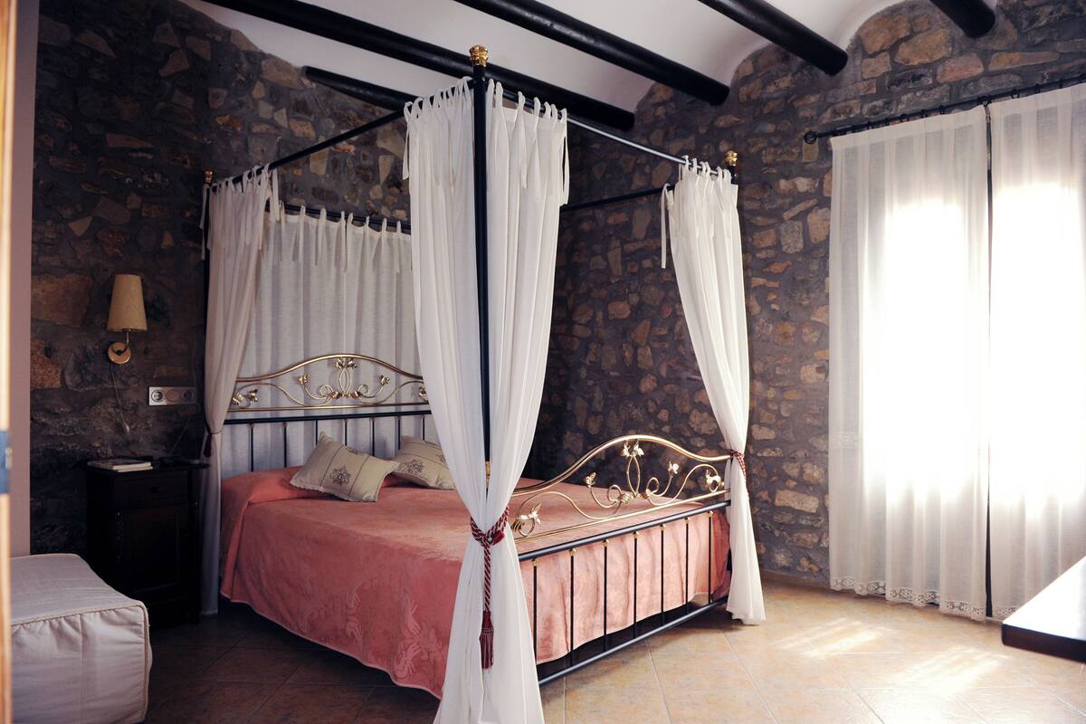 A four-poster bed with white drapes, a stone wall, and a nightstand with a lamp.