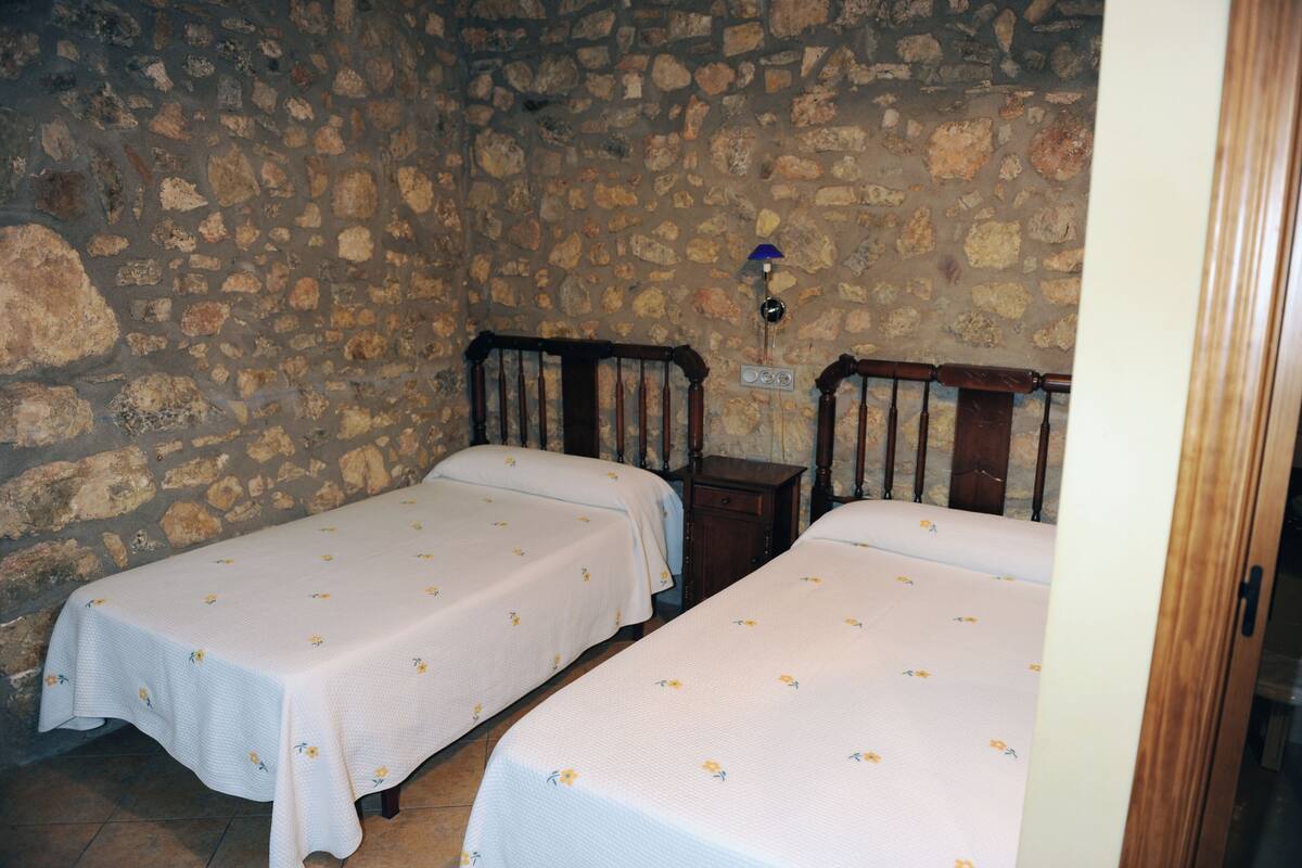 A room with two wooden beds, each with a white bedspread featuring a small floral pattern. The walls are made of stone, and there is a small wooden nightstand between the beds.