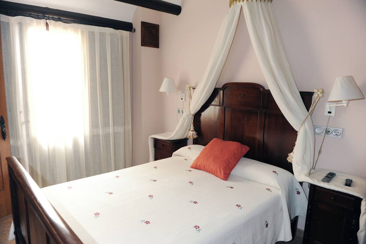 A neatly made bed with a floral bedspread, a wooden headboard, and a canopy. There are two bedside tables with lamps and a nightstand with a lamp.