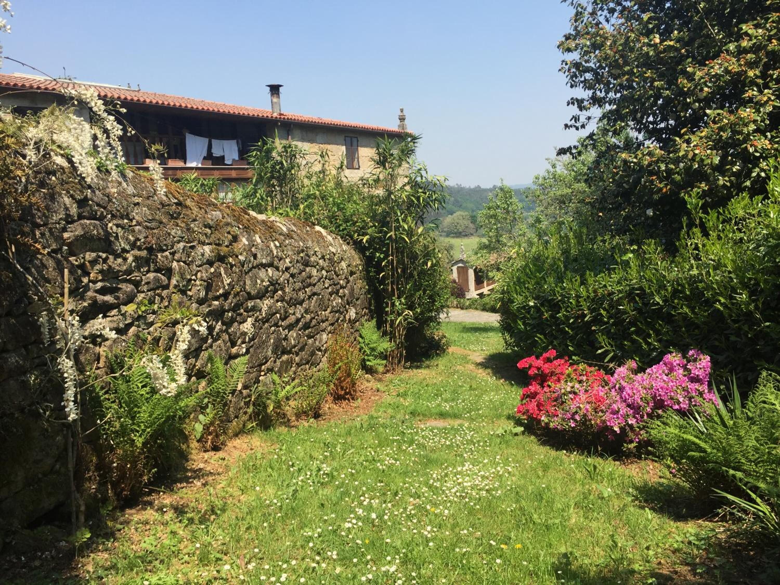 A stone wall, a stone building with a red-tiled roof, and a garden with flowers and greenery.