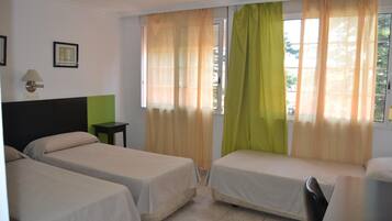 Habitación de hotel con dos camas individuales, una mesita pequeña, una silla y una ventana con cortinas translúcidas.