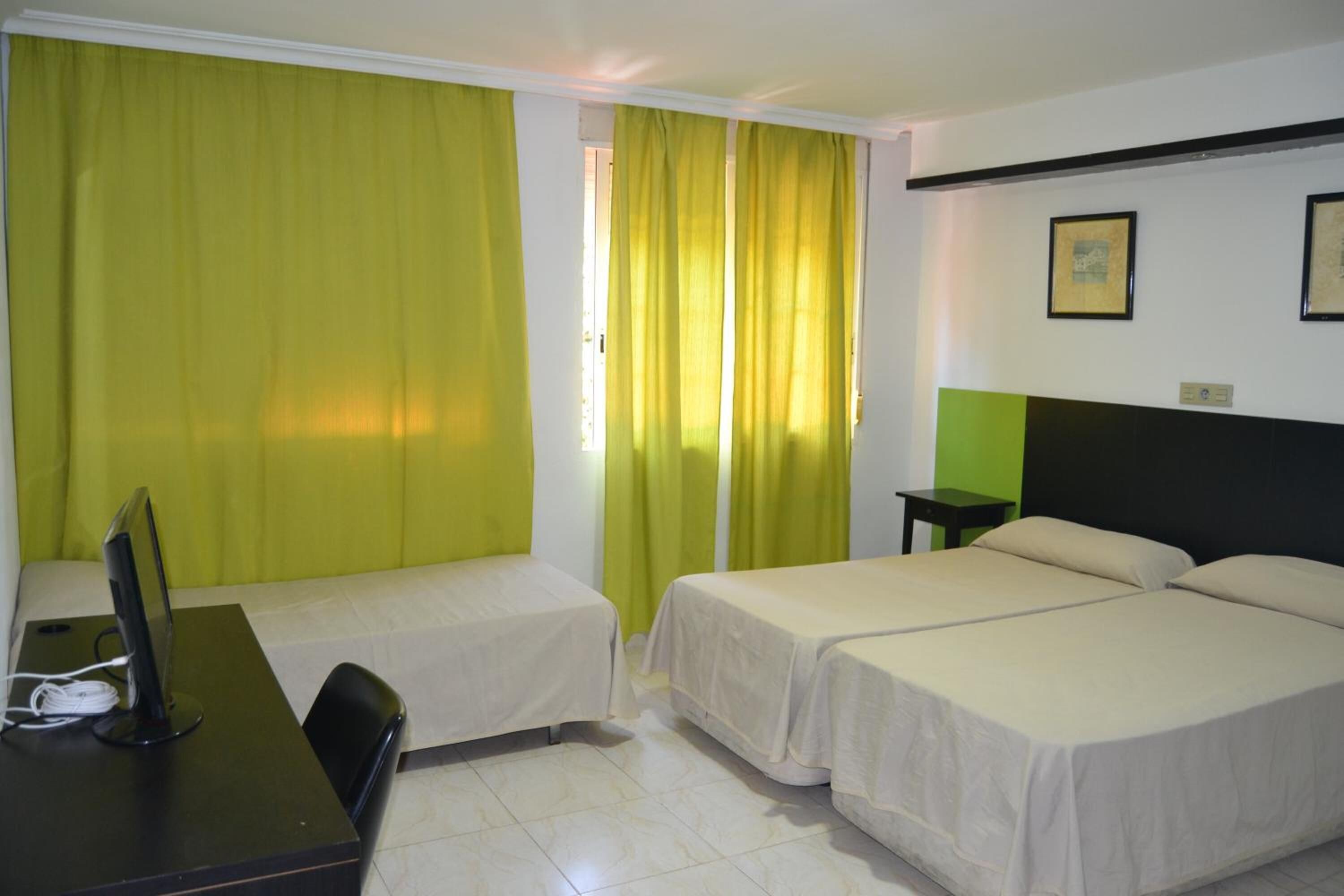 Habitación de hotel con dos camas, un escritorio con computadora y una ventana con cortinas verdes.