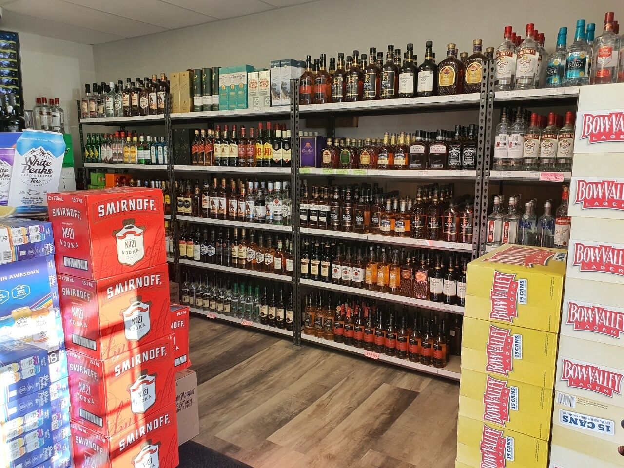 A liquor store with shelves stocked with various bottles and boxes of alcohol, including brands like Smirnoff and Bow Valley Lager.