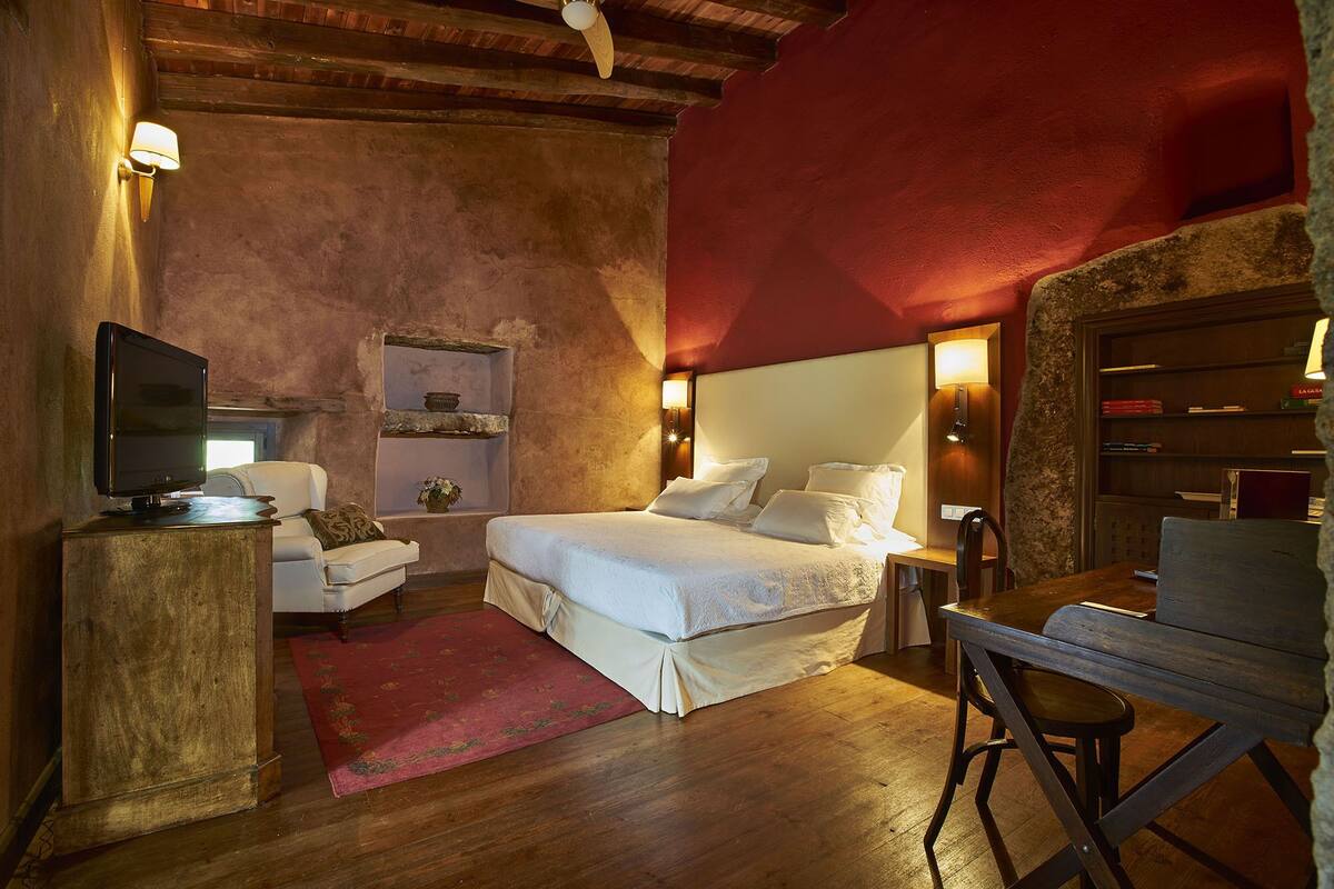 A cozy bedroom with a wooden ceiling, a bed with white linens, a desk with a chair, and a television on a wooden stand.